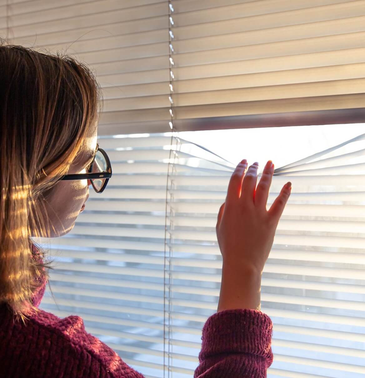 Frau schaut durch Jalousien und lässt Sonnenlicht in den Wohnraum