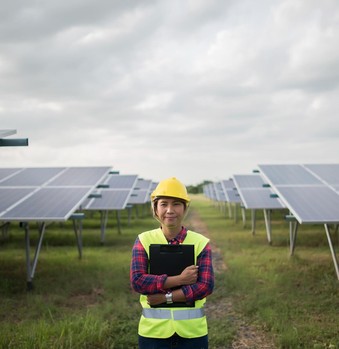 Elektrikerin mit Schutzhelm und Klemmbrett steht vor Reihen von Solarmodulen